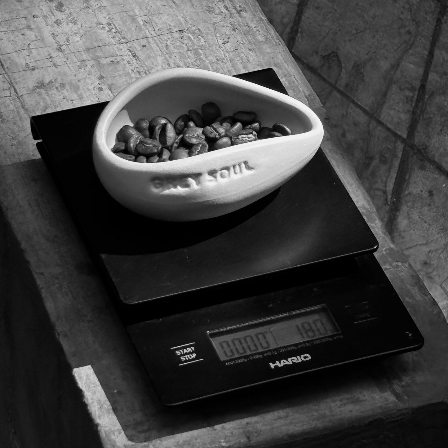 A white stoneware matte coffee bean doser with the text 'GREAT SOUL' embossed on it, placed on a black scale with a bowl of coffee beans behind it.